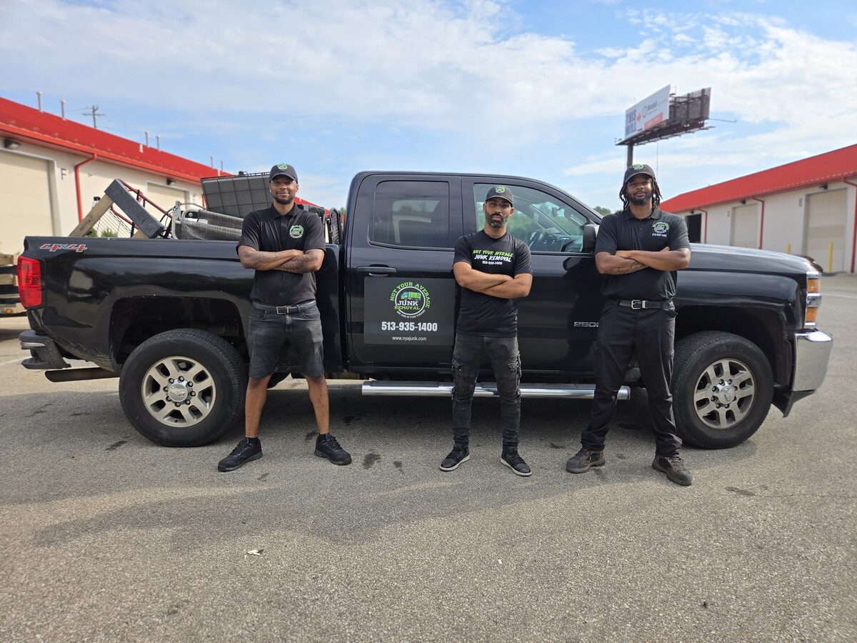 Three NYA Junk Removal crew members with arms folded in front of their branded pickup truck, which carries the company logo and phone number on the door.