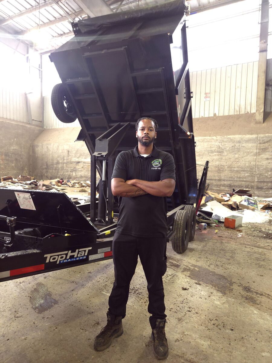 An NYA Junk Removal crew member standing arms folded in front of the raised dump trailer as it empties a load.
