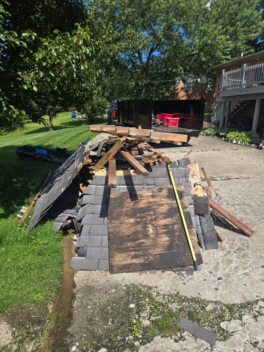 A pile of torn-out deck boards, joists, roofing shingles and plywood in a driveway, with the NYA dump trailer parked behind it.