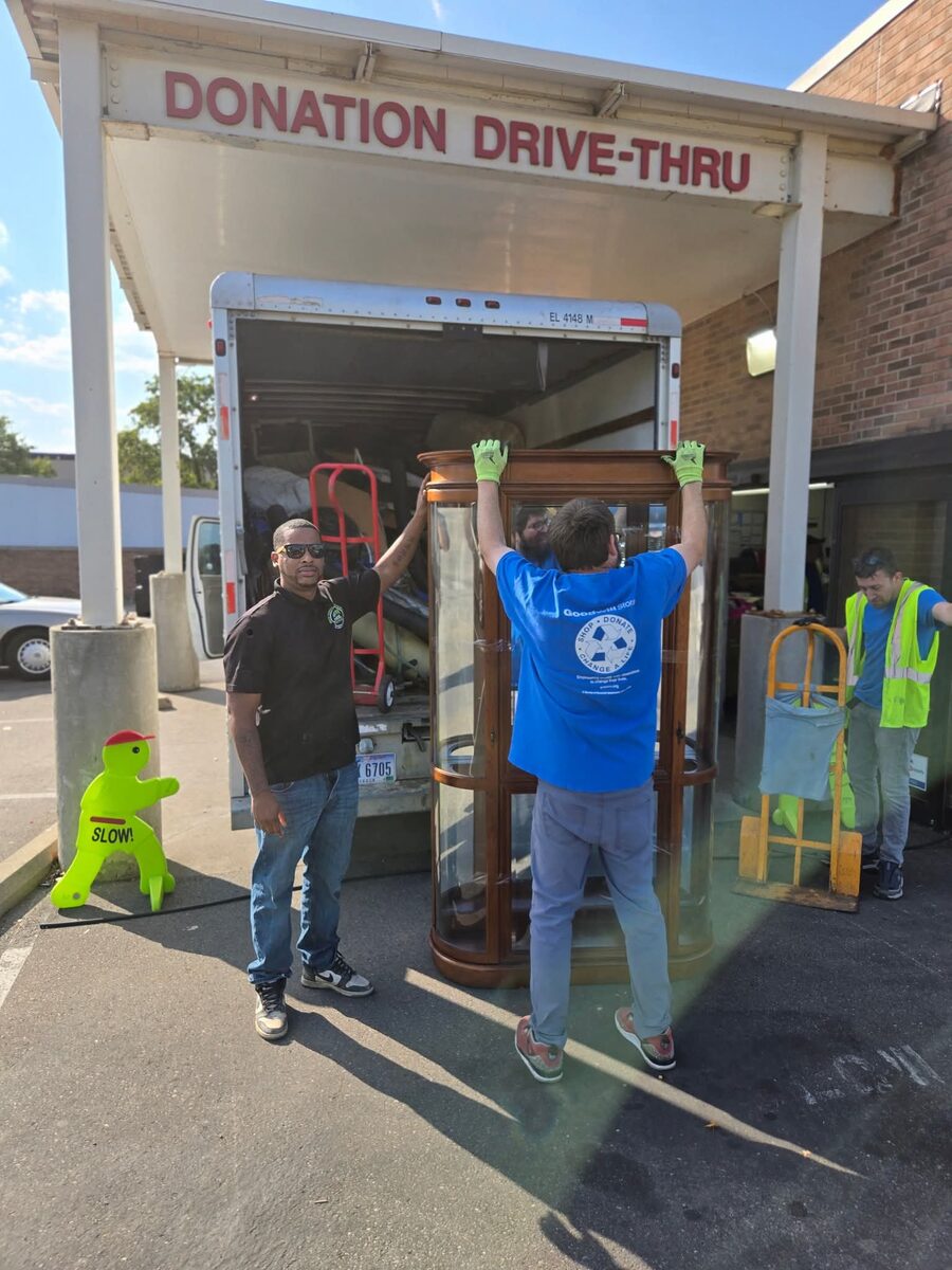 NYA crew unloading a tall wooden curio cabinet from the box truck under a donation drive-thru canopy.