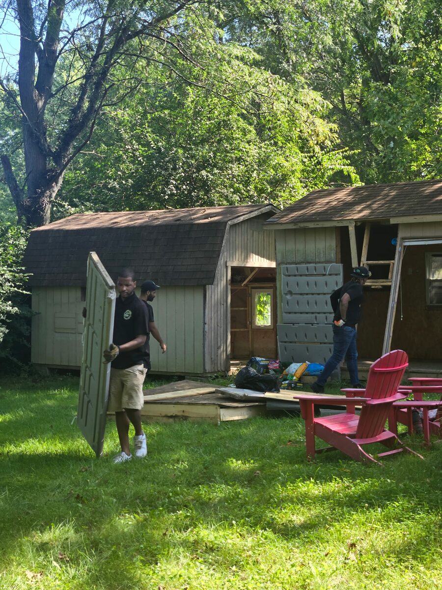 NYA crew taking down a pair of backyard sheds in Middletown, carrying out a door panel past red lawn chairs.