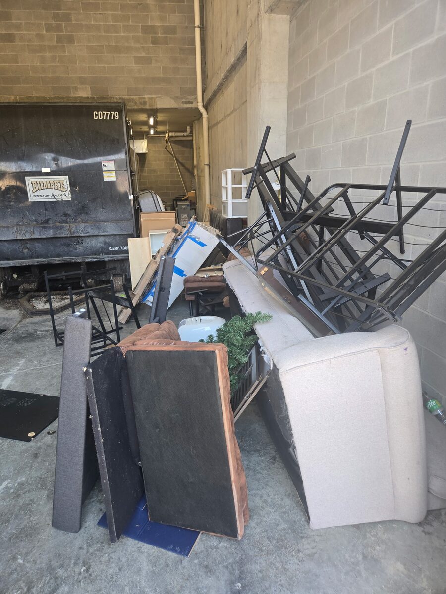 A building’s dumpster bay crowded with a couch, chairs, cushions, metal chair frames, boxes and a small artificial tree.