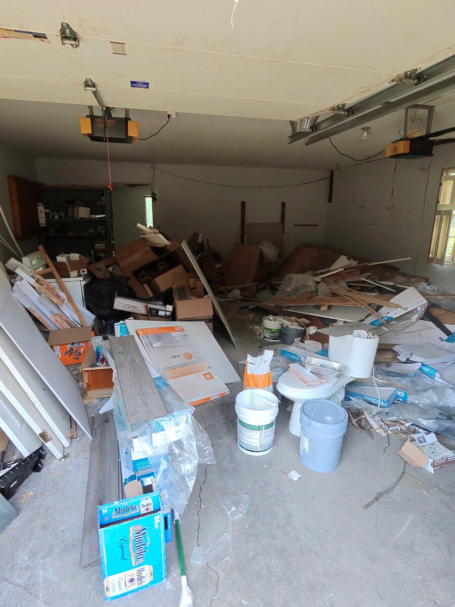 A two-car garage packed with renovation debris — broken drywall, offcut lumber, paint buckets and flooring boxes covering the floor.