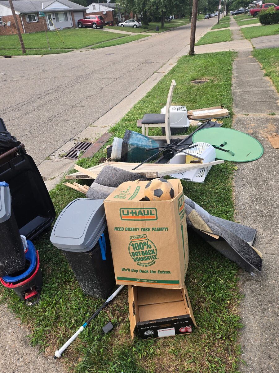 A residential tree lawn piled with bins, a folding chair, rolled carpet and boxes set out for curbside pickup.