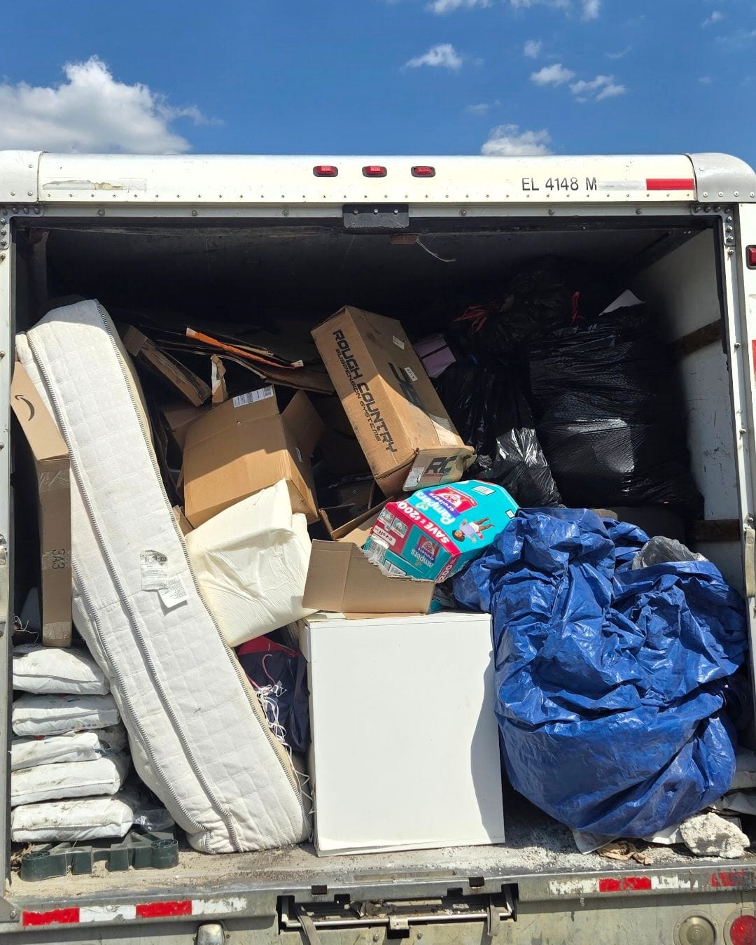 The open back of the NYA box truck packed with a mattress, box springs, cardboard boxes, a small white appliance and bagged household items.