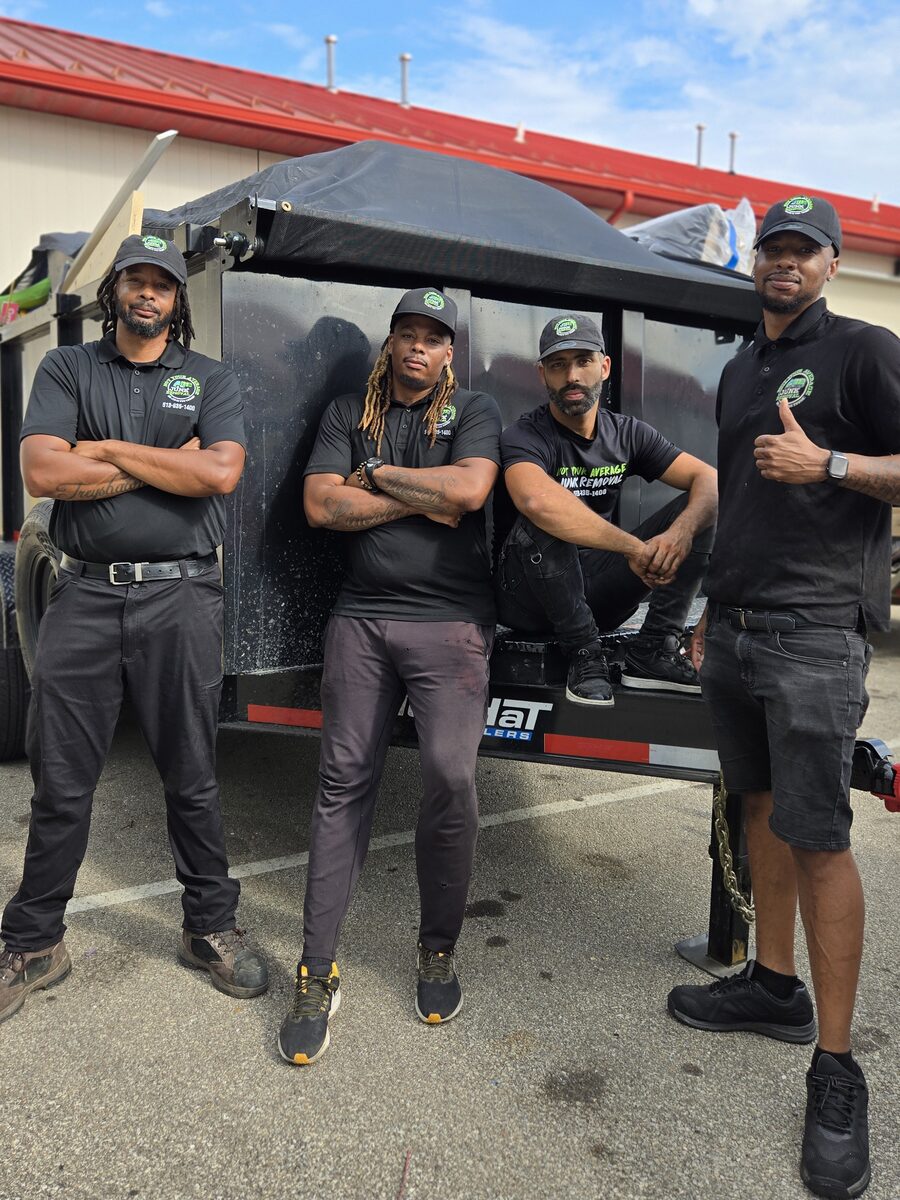 Four NYA Junk Removal crew members leaning against their black high-wall dump trailer, one giving a thumbs up.