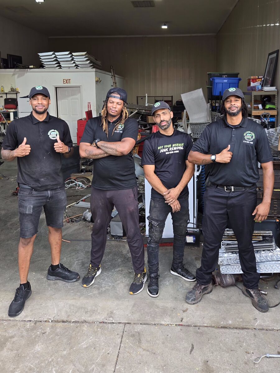Four NYA Junk Removal crew members giving a thumbs up in front of a warehouse bay stacked with scrap, appliances and hauled items.