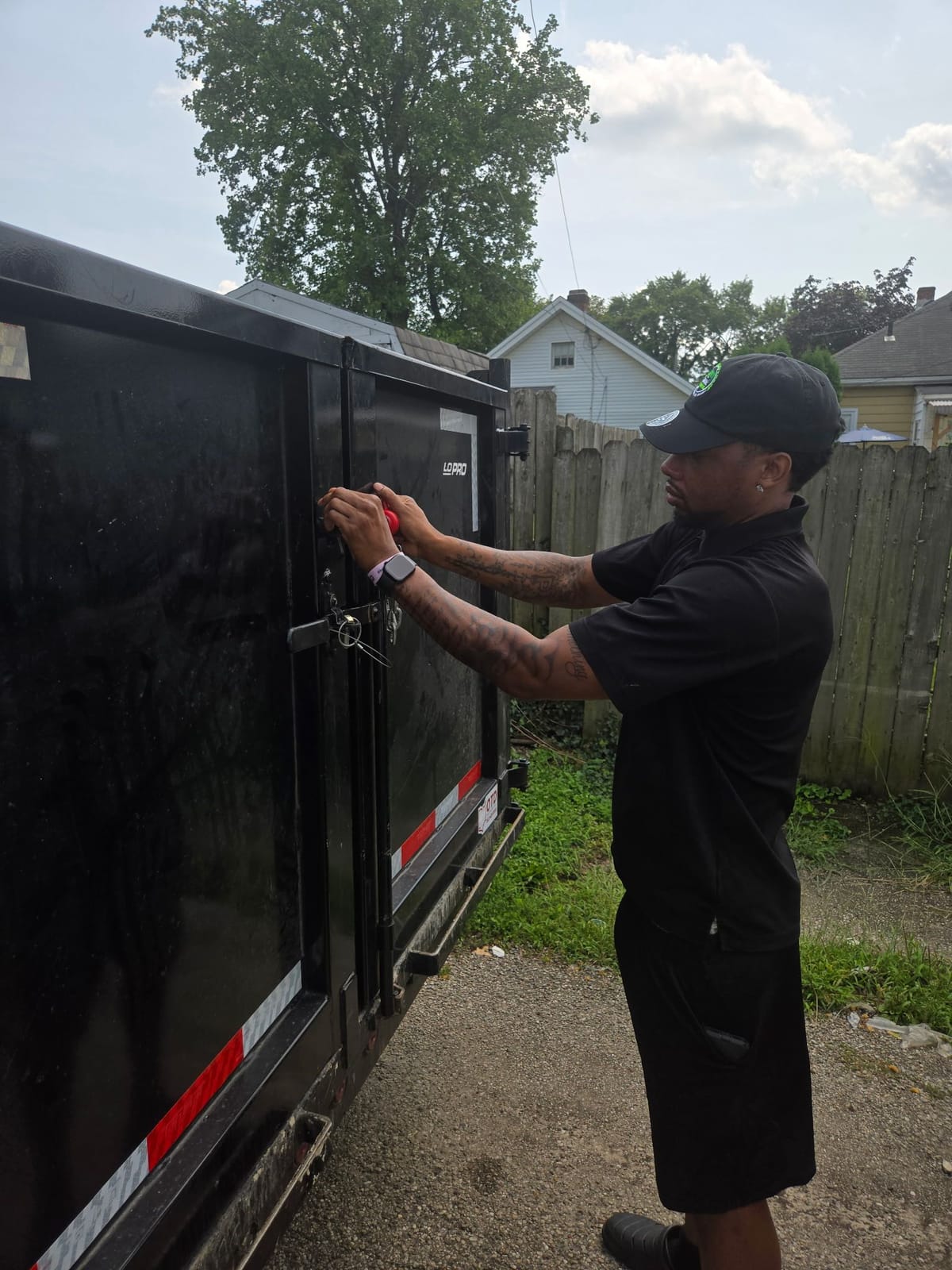 An NYA Junk Removal crew member in a branded cap and polo unlatching the rear gate of the black dump trailer at a residential job.
