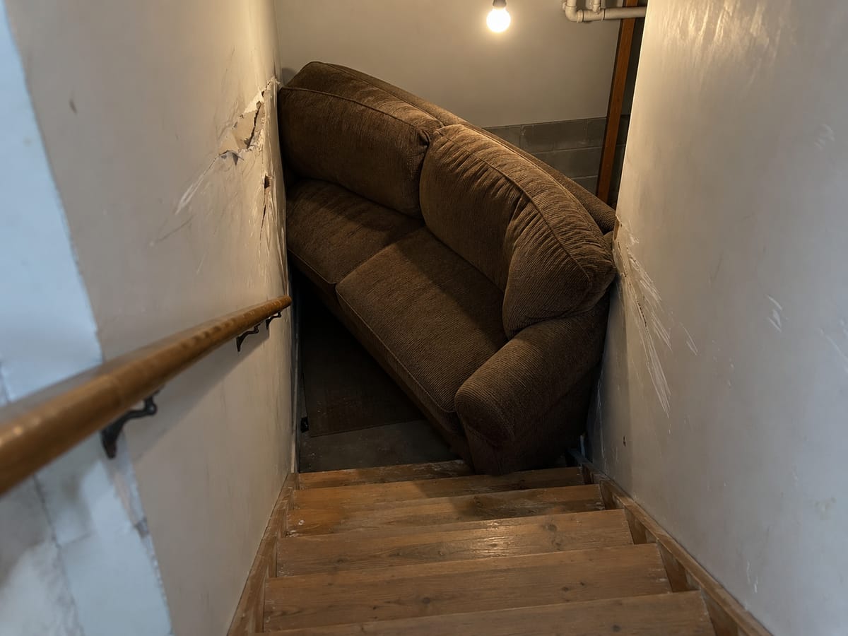 A brown sofa wedged at an angle on a narrow basement stairwell, scraping the wall beside the handrail.